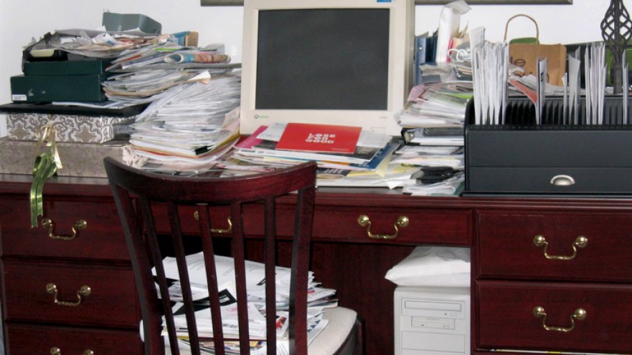 Desk with computer, piled high with stacks of papers.